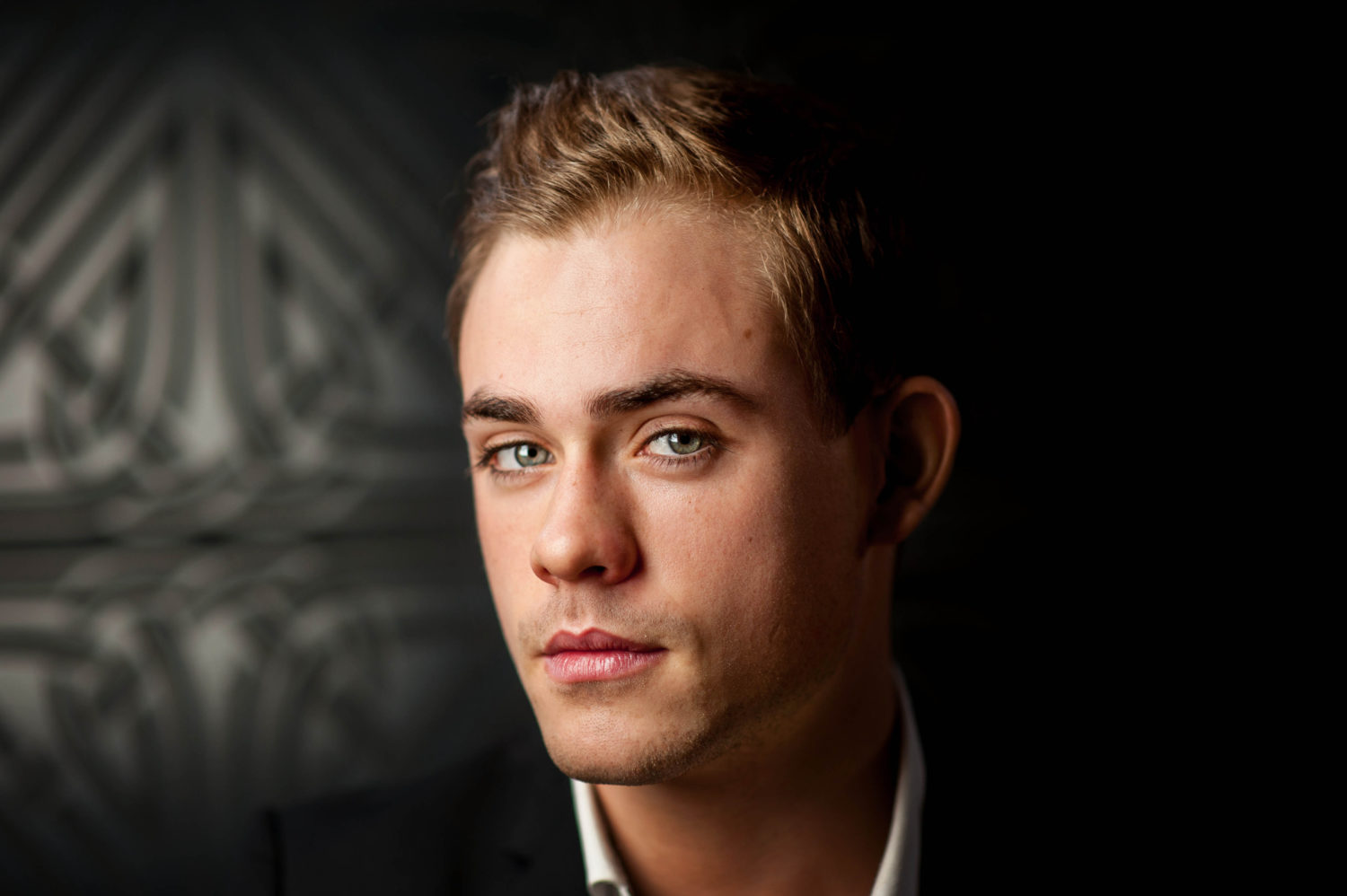 A young man with blond hair and blue eyes wearing a black suit